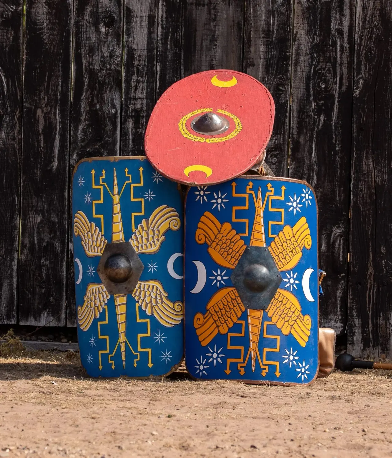 Colourful painted wooden shields depicting Roman military insignia, displayed against weathered dark wooden planking.