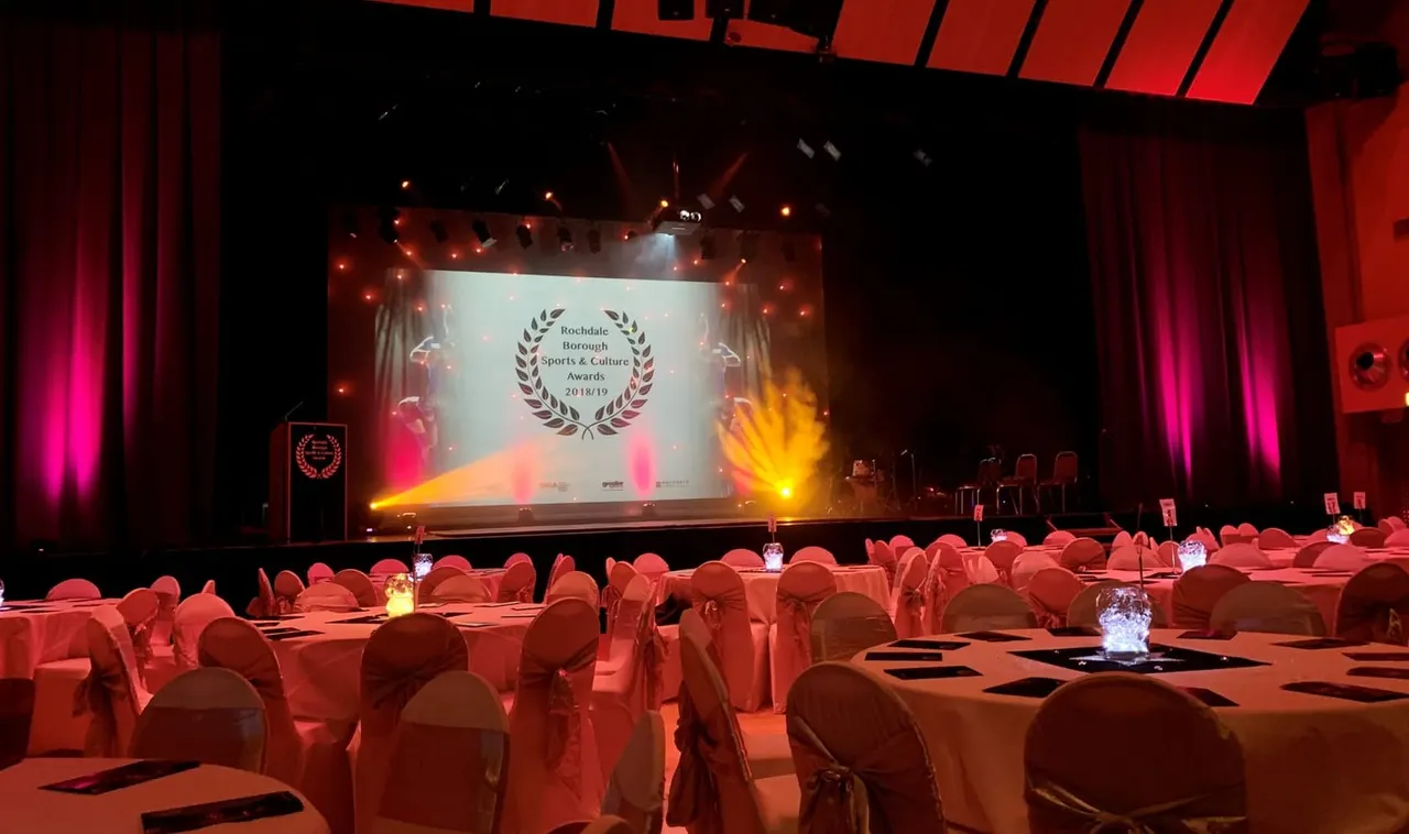 Gala event hall with tables and chairs arranged facing a large lit stage displaying an awards ceremony presentation.