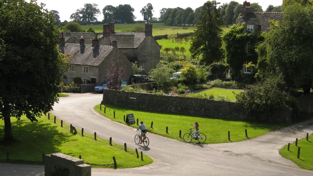 Two cyclists riding on a curved path through a historic stone village with countryside fields beyond.