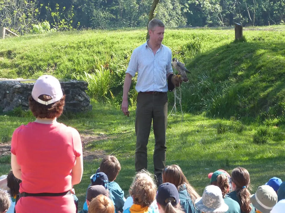 Man in blue shirt holding a bird of prey demonstrates to a seated group of children on a grassy field.
