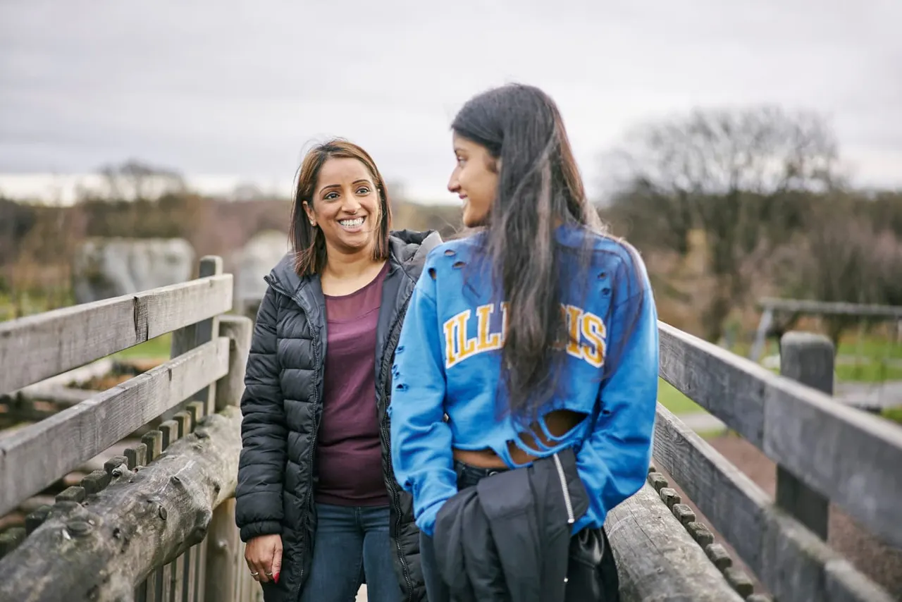Two young women smile whilst standing on a wooden bridge overlooking countryside with bare trees and fields.
