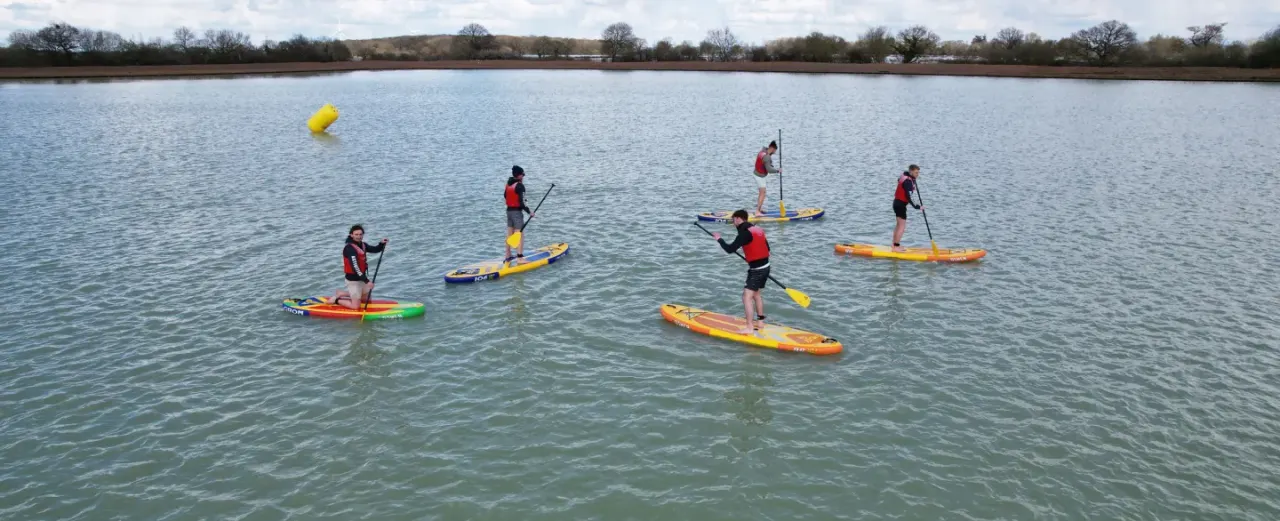 Group of people stand-up paddle boarding on a lake with yellow buoys marking a course.