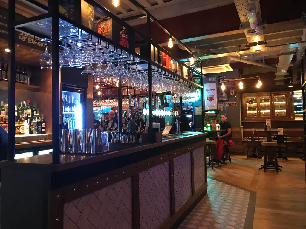 Interior of a pub with a long bar counter, hanging glassware, bottles on shelves, and customers seated at tables.