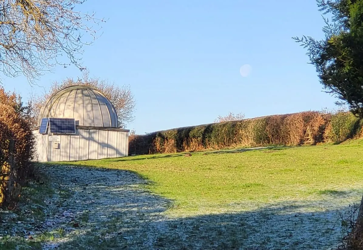 Observatory dome building with solar panel in green field beside autumn hedgerows on clear day.