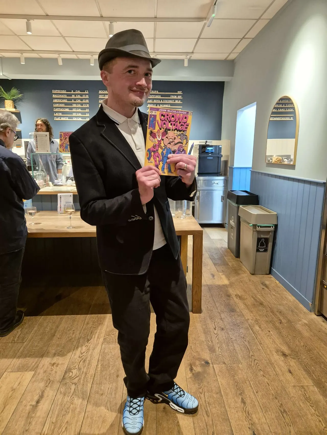 A man in a black suit and fedora hat holds a colourful comic book whilst standing in a café with menu boards and other customers visible.
