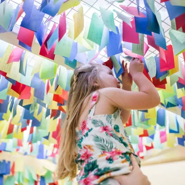 Young child reaching up towards colourful hanging paper decorations in an interactive indoor installation.