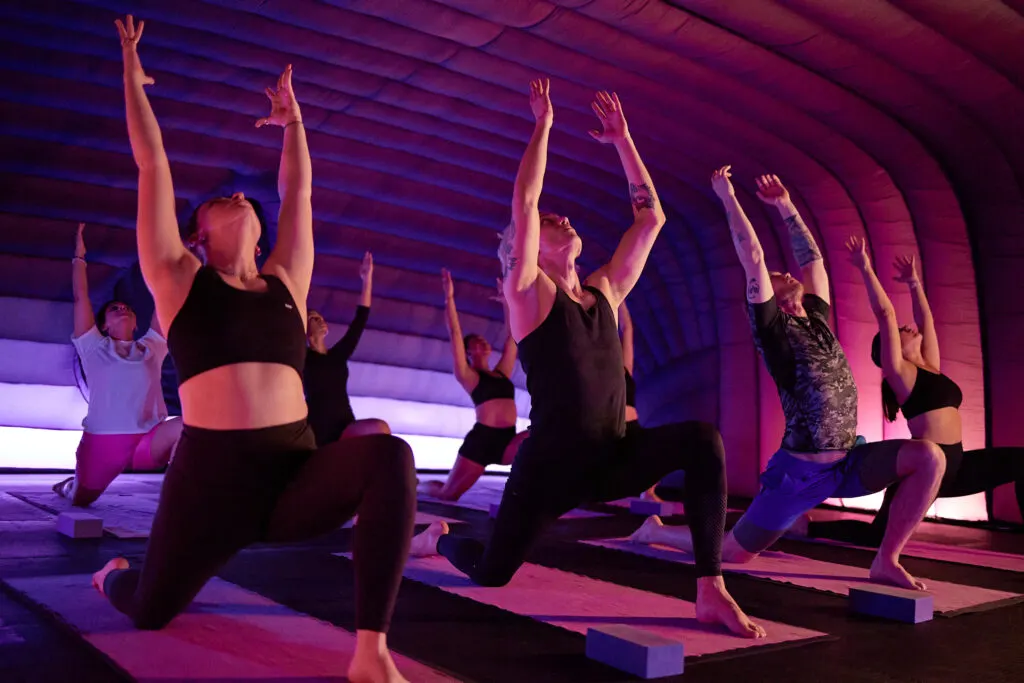 Group of people in a yoga class performing a lunge pose in a purple-lit studio.