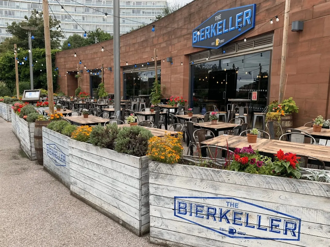 Outdoor beer hall terrace with wooden tables, metal chairs, colourful planted beds and hanging string lights beside a brick building.