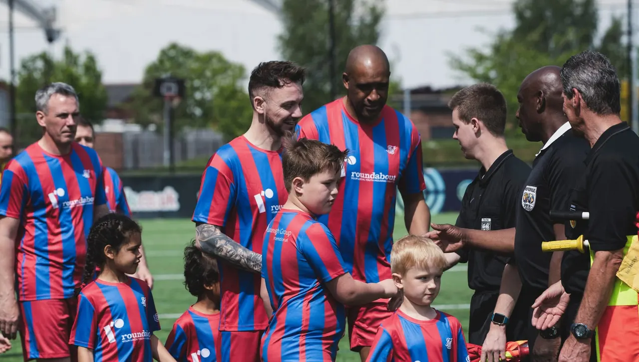 Football players and young children in red and blue striped kits with match officials on a football pitch.