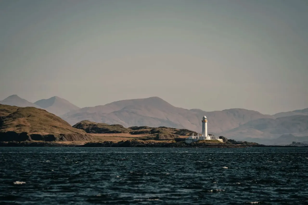 White lighthouse on rocky coastline with mountains in distant haze, viewed across dark sea.
