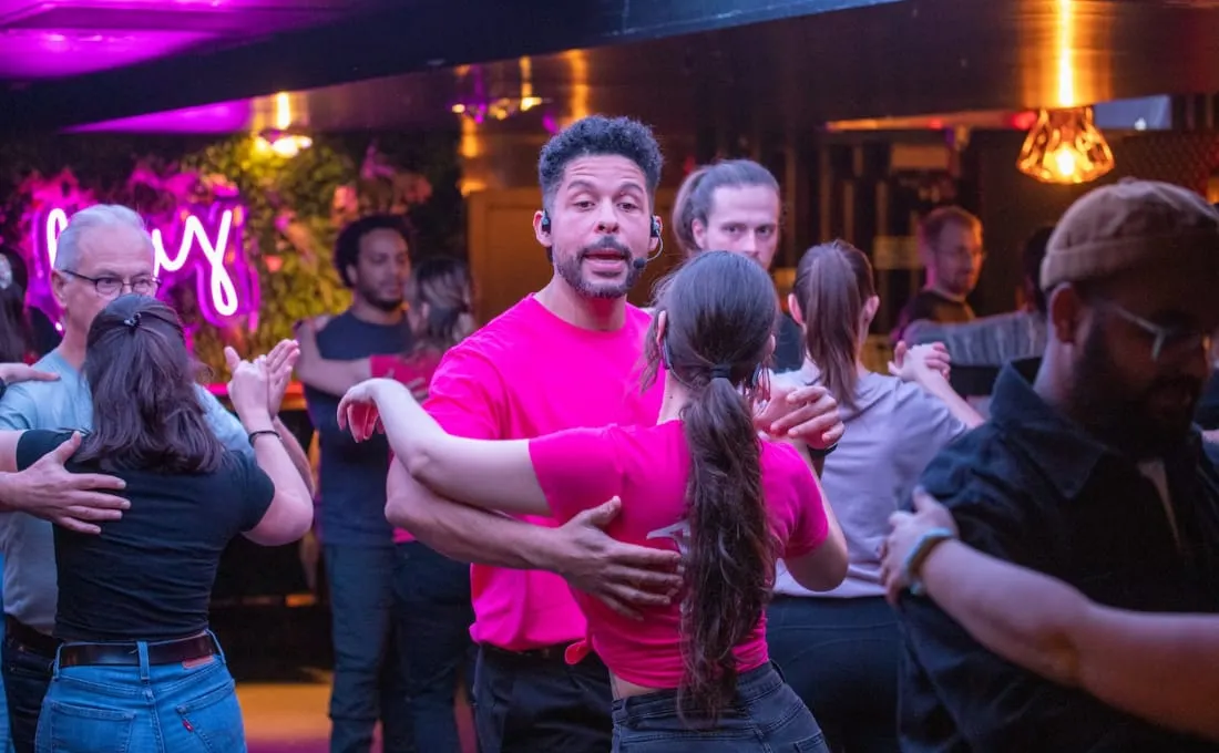 Group of people dancing together in a neon-lit venue with magenta and gold lighting and decorative plants.