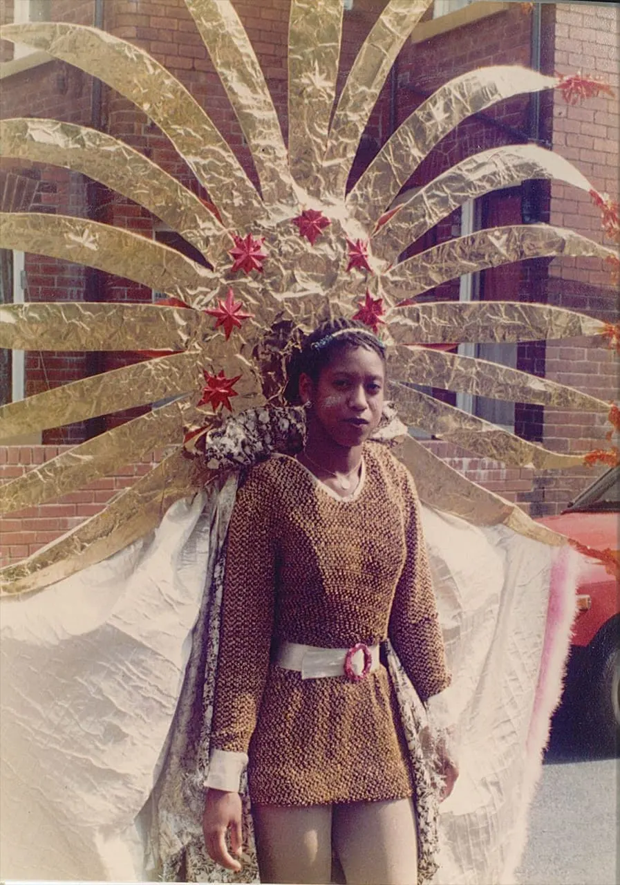 Child wearing an elaborate golden and white feathered headdress and cape with red star decorations stands in front of a brick building.