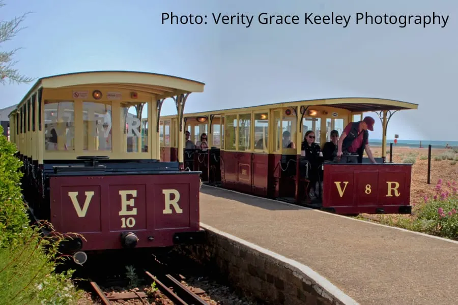 A vintage open-air electric railway train with cream and maroon carriages stops at a seaside station with passengers boarding.