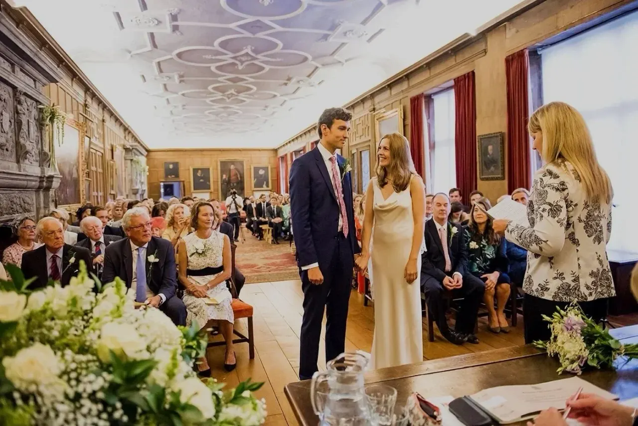 A couple in wedding attire stand in an ornate golden hall during a formal ceremony, with seated guests lining both sides and floral arrangements in foreground.