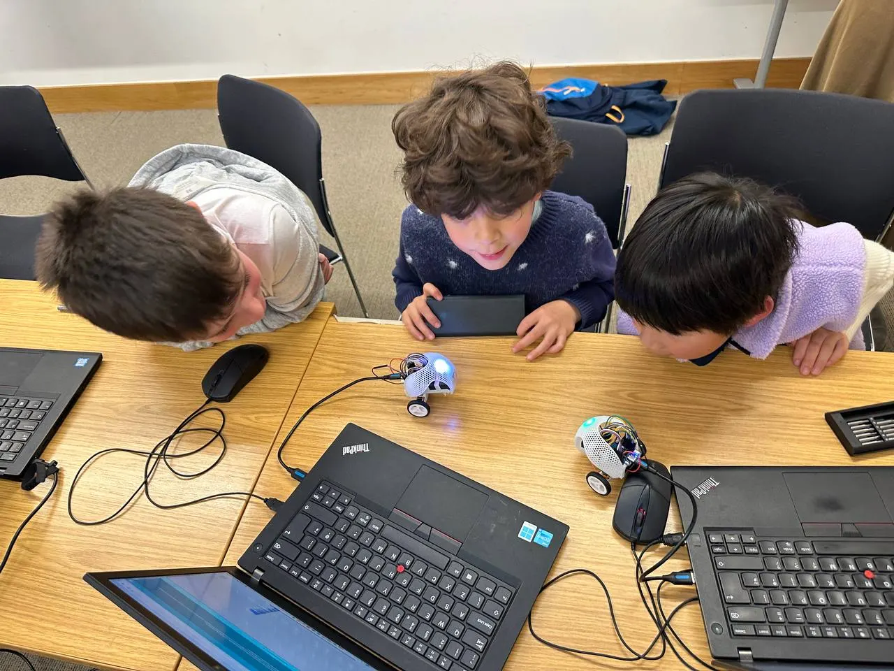 Three children engaged in a coding or robotics workshop, using tablets and laptops at a wooden table with small robotic devices.