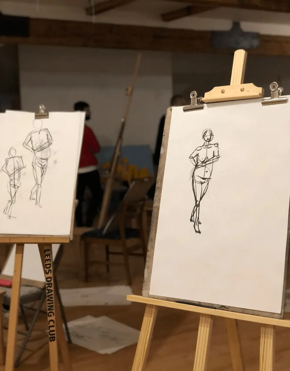 Figure sketches on easels during a life drawing class with a model in the background.