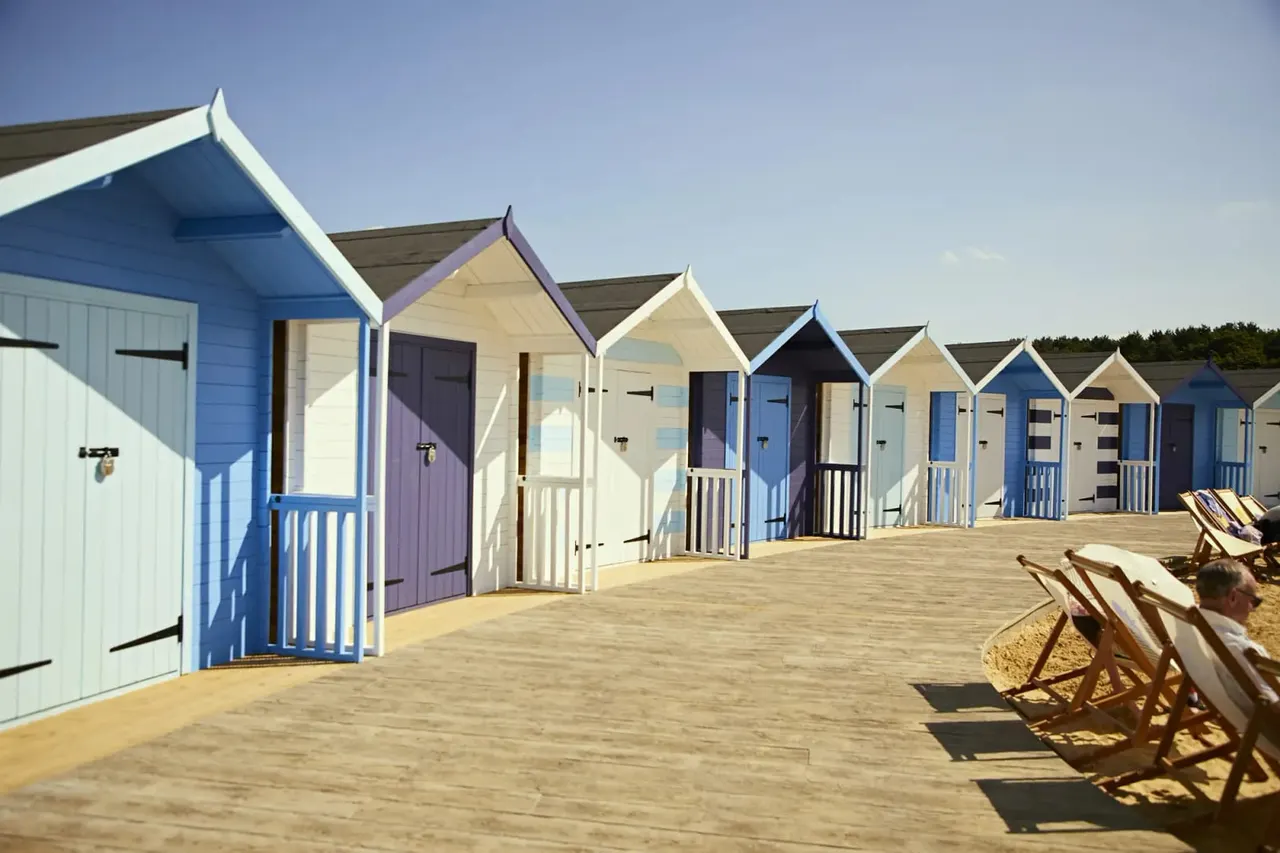 Row of colourful beach huts on sand with deckchairs in foreground under clear blue sky.