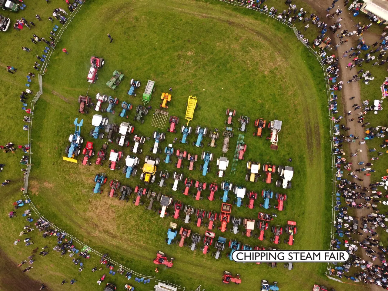 Aerial view of a steam fair with dozens of vintage tractors and farm machinery displayed in rows on green grass, surrounded by visitors.