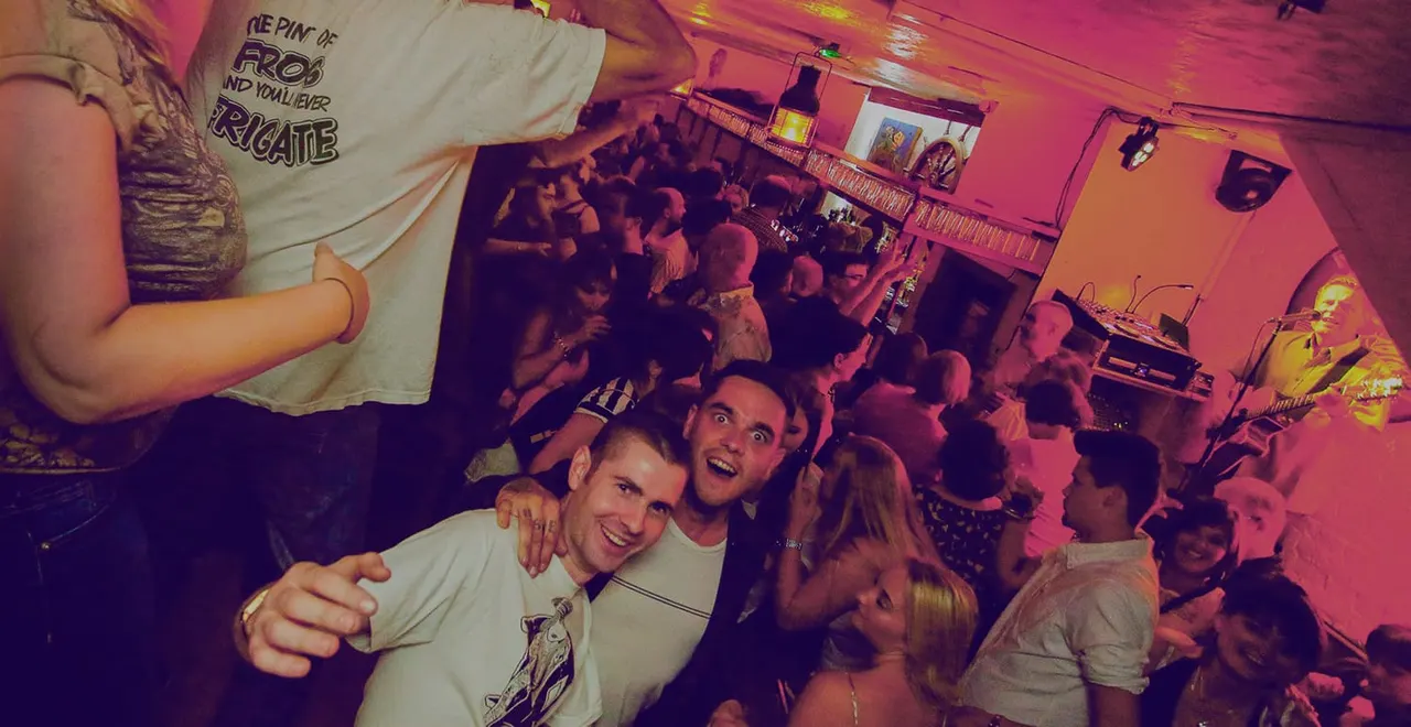 Crowds of people dancing and socialising in a dimly lit nightclub with pink and red lighting and a bar in the background.