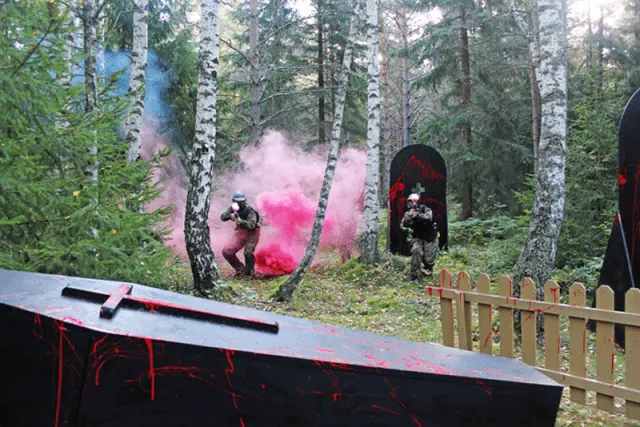 Players in combat gear fire paintball guns surrounded by pink powder clouds in a woodland arena.