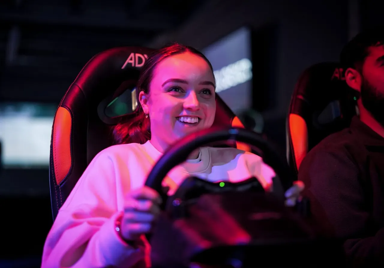 Young person smiling while holding a steering wheel in a racing simulator, illuminated by red and pink neon lighting.