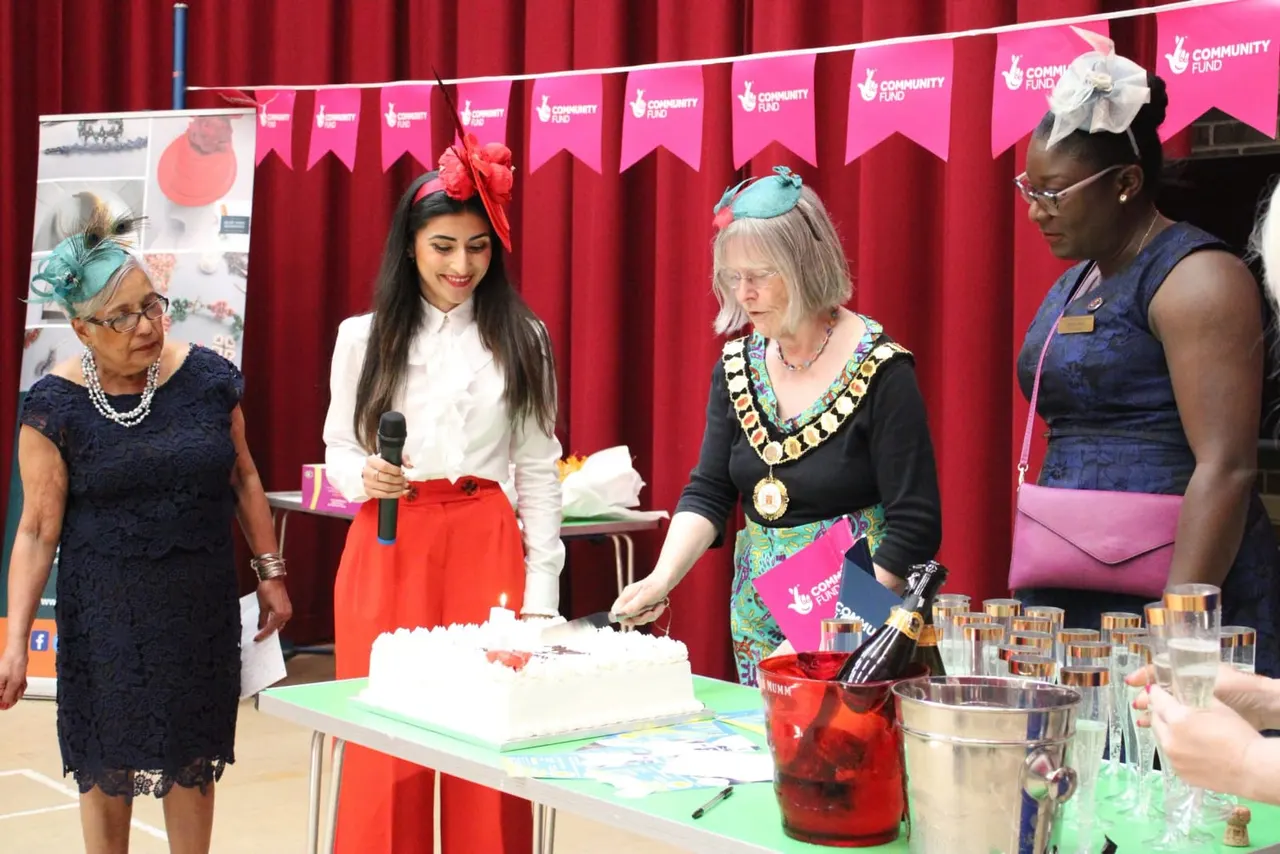 Four women at a formal event cutting a white cake, with red curtains and pink Community Fund bunting behind them.