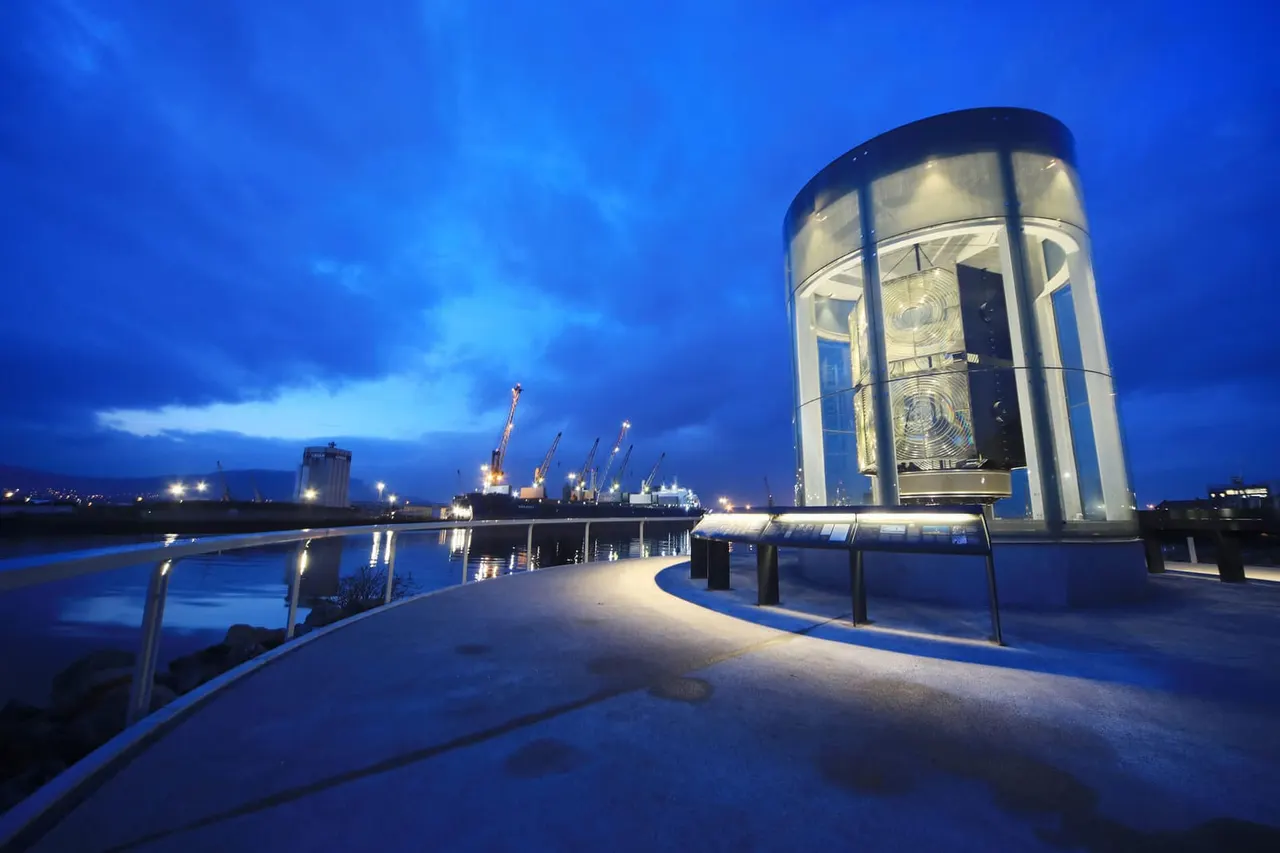 Illuminated cylindrical lighthouse structure on waterfront promenade at dusk, with industrial port and cranes visible across the water.