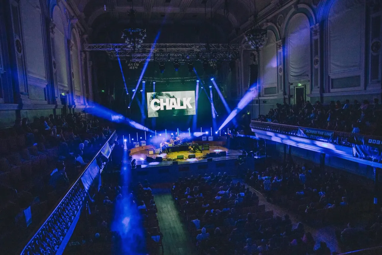 Large concert hall with crowd seated around a stage lit by blue lighting beams, with "CHALK" displayed on screen above.