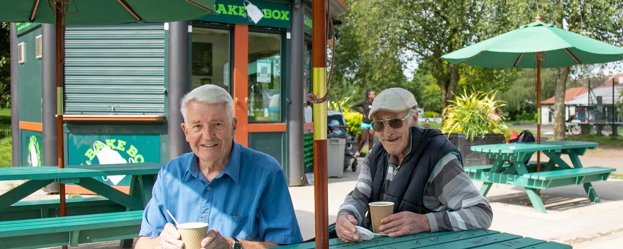 Two older men sitting at a green picnic table outdoors holding drinks, with a takeaway kiosk and green umbrellas behind them.