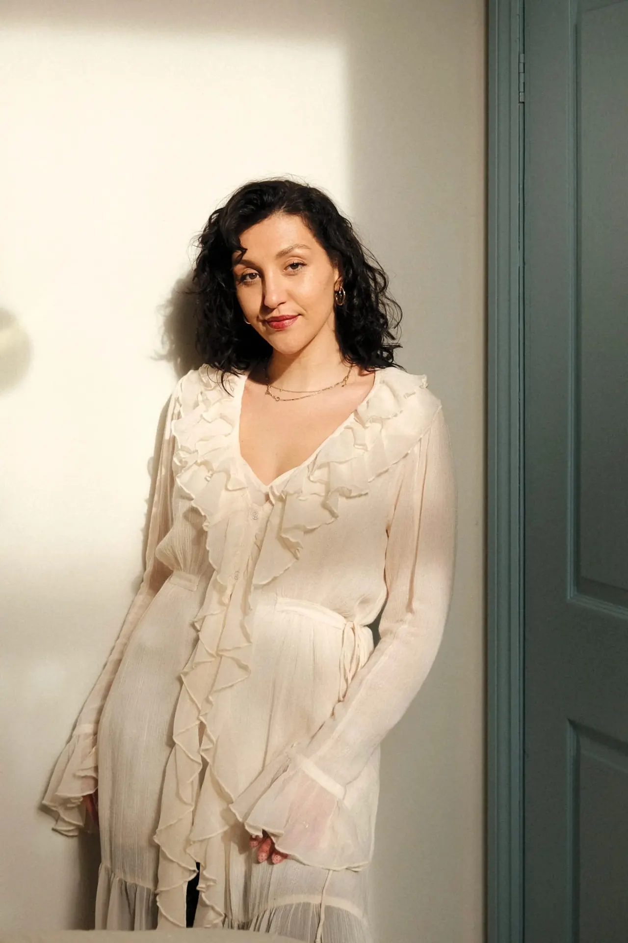 Woman with black wavy hair wearing a cream ruffled blouse standing against a white wall.