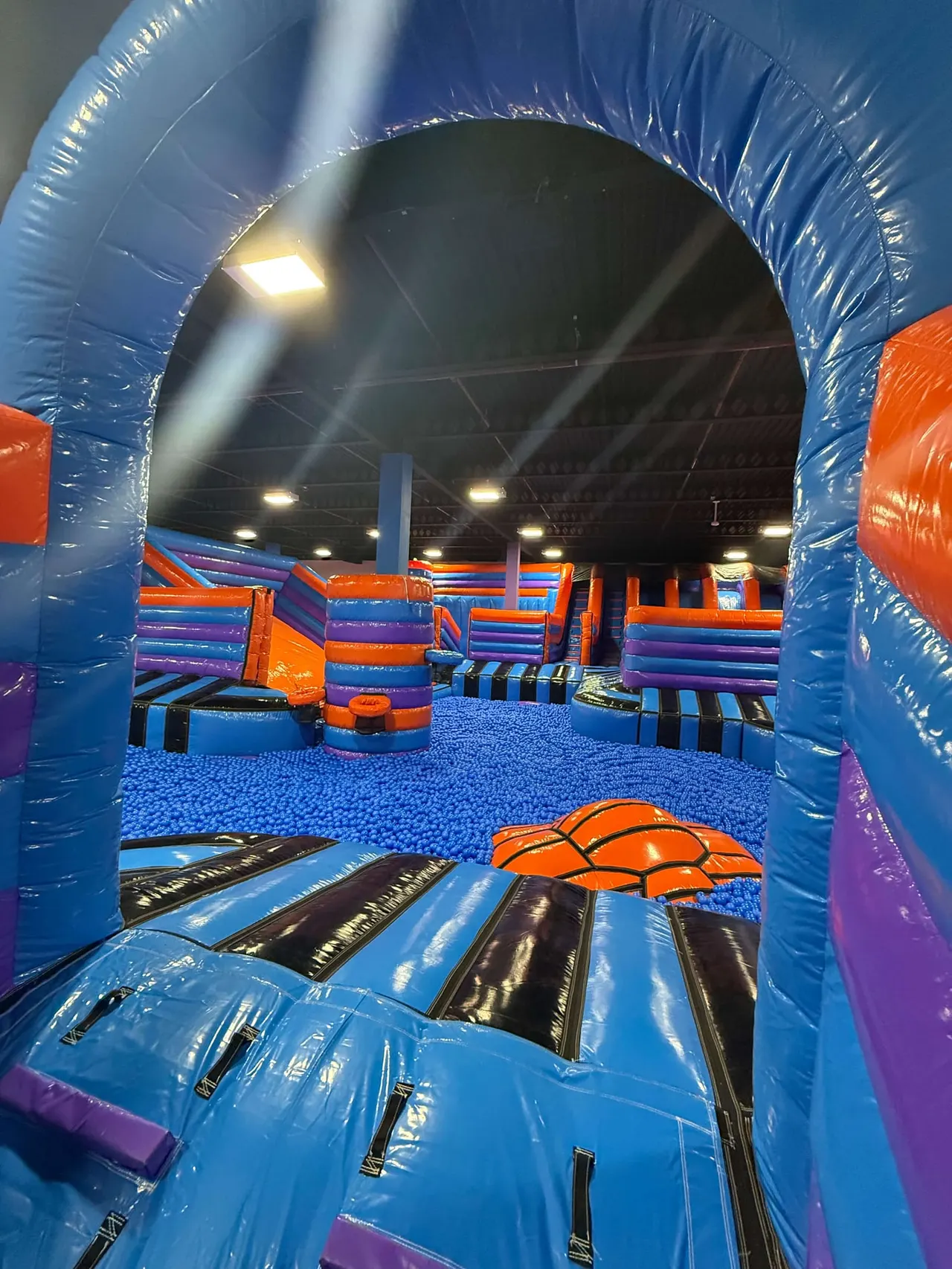 Interior view through a blue inflatable tunnel of a colourful indoor play arena with bouncy castle equipment and obstacle courses.