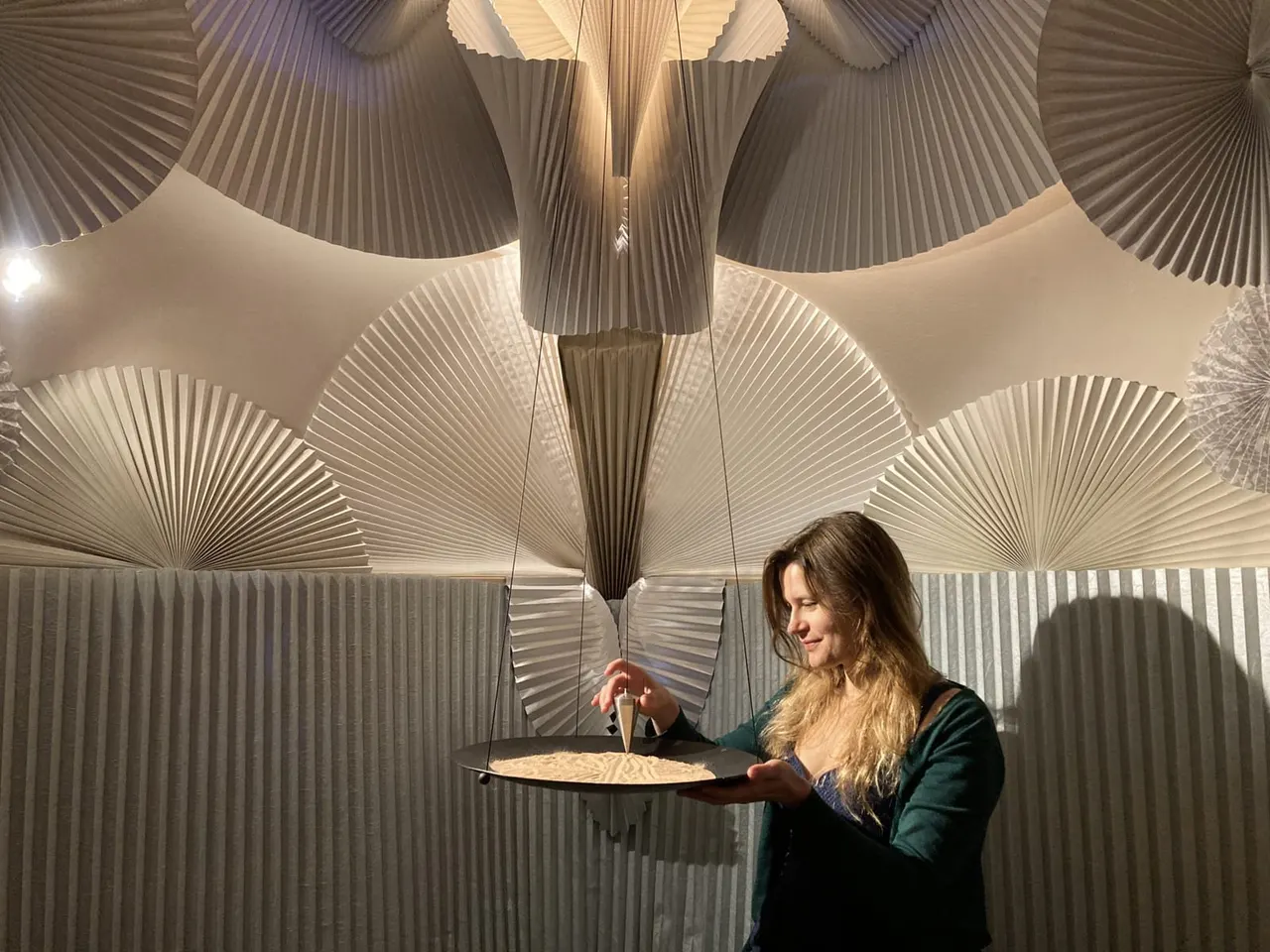 Woman holding a plate of food in an illuminated gallery space with pleated sculptural wall panels.