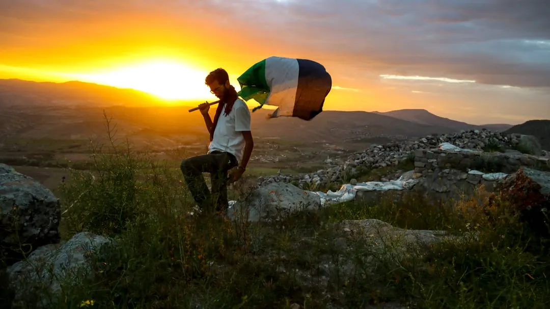 Person flying a kite on a rocky hillside at sunset overlooking a valley with mountains and settlements.