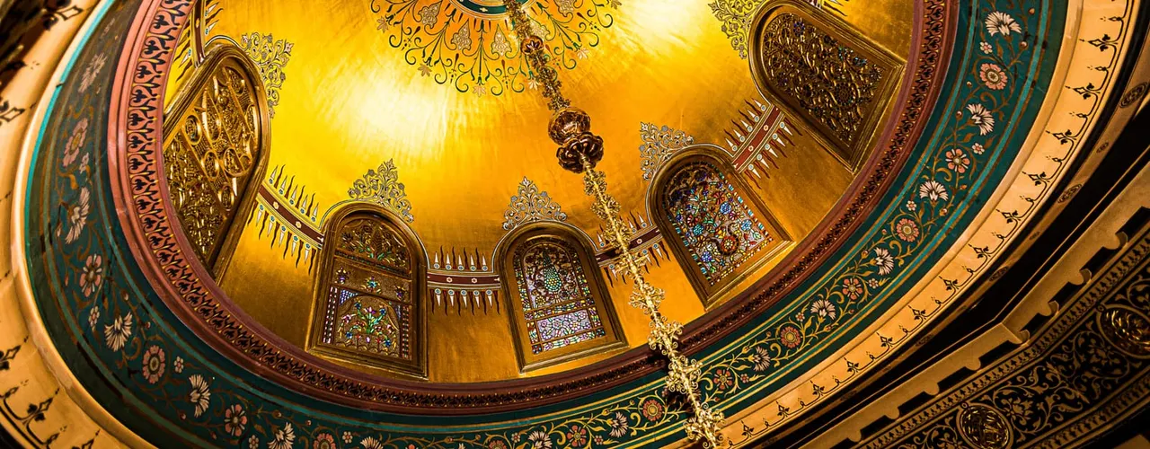 Ornate gilt dome ceiling with jewel-toned decorative panels and intricate carved details in an interior architectural space.