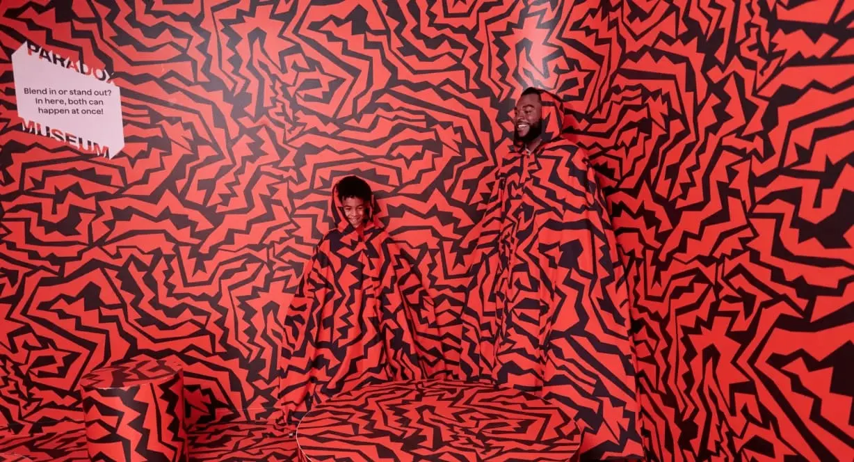 Two visitors pose together in front of a large red and black abstract patterned wall installation at a museum.