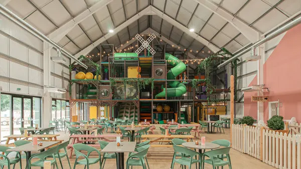 Indoor farm shop café with green tables and chairs beneath a high vaulted ceiling decorated with hanging plants and colourful balloons.