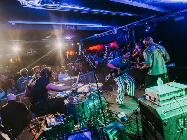 Live band performs on stage to a crowded audience in a dimly lit indoor venue with blue and green lighting.