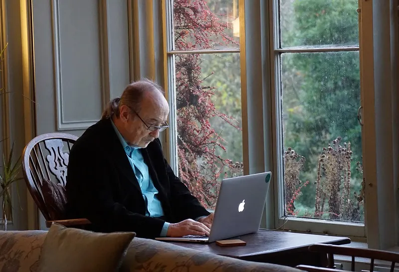 Man in business attire working on laptop at a table by large windows overlooking autumn woodland.