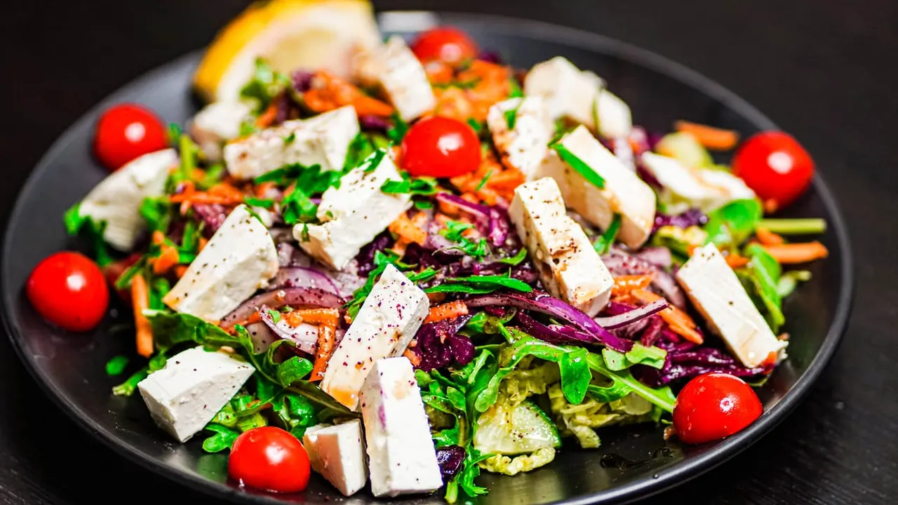 Colourful salad with mixed greens, sliced white cheese, roasted vegetables, cherry tomatoes and fresh herbs on a dark plate.