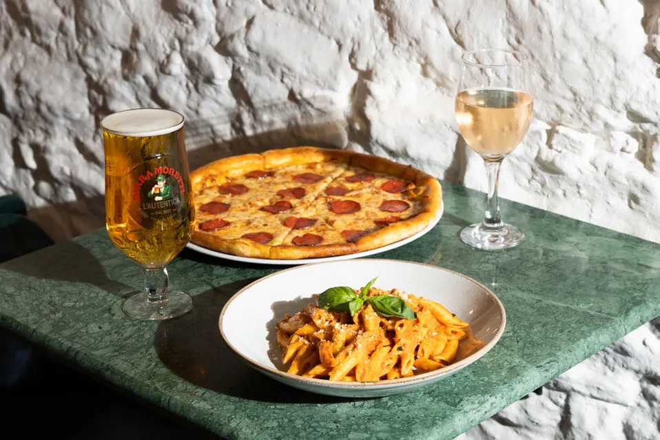 A table set against a white stone wall displays a pepperoni pizza, a bowl of dressed fries with basil, a beer and a glass of white wine.