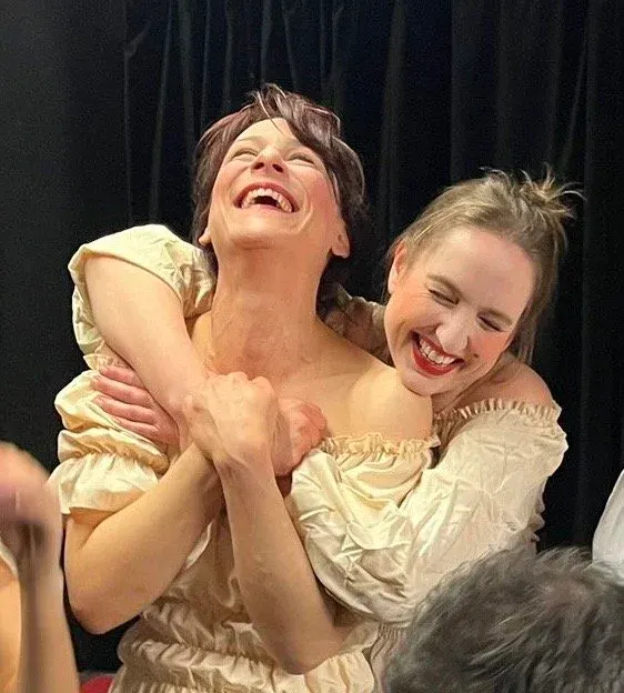 Two performers in cream-coloured period costumes laugh together on stage during a theatrical production.