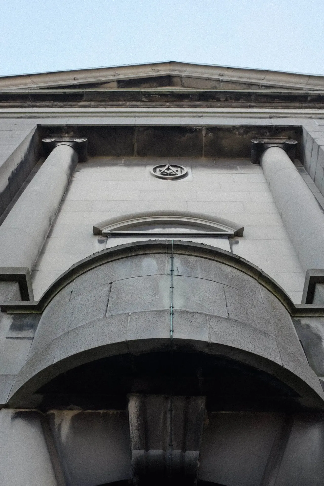 Upward view of a classical stone building façade featuring fluted columns, a large arched doorway, and decorative stonework against a clear sky.