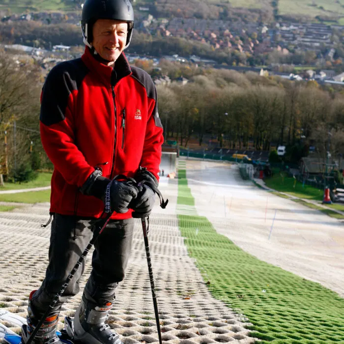 A skier in a red and black jacket and helmet stands on a dry ski slope with a suburban valley landscape visible behind.
