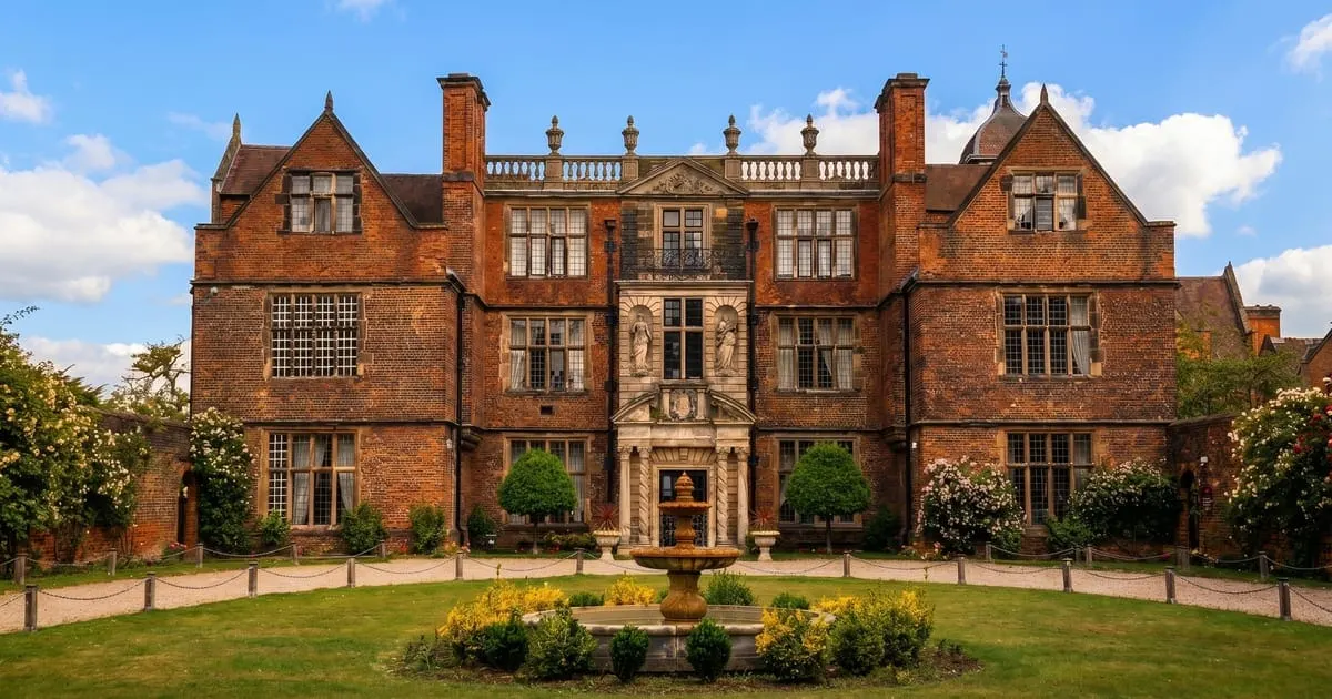 Large red-brick Jacobean mansion with manicured gardens, topiary, and a central fountain under blue sky.