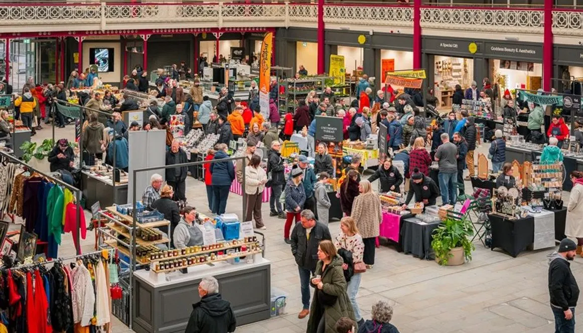 Busy indoor market hall with numerous shoppers browsing stalls selling clothing, crafts and food under ornate Victorian ceiling.