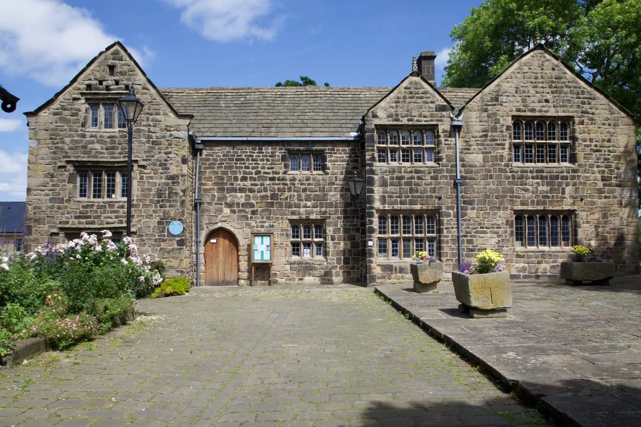 Stone manor house with gabled roofs, multi-paned windows, and flowering plants in front courtyard on sunny day.