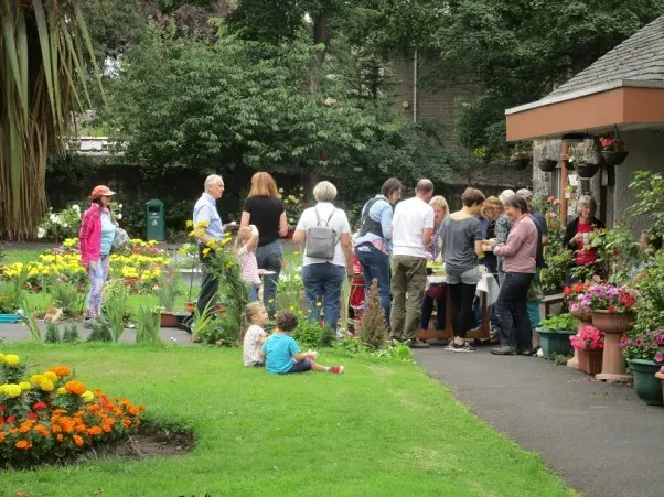Visitors of various ages exploring a garden with flower beds and potted plants near a brick building and woodland setting.