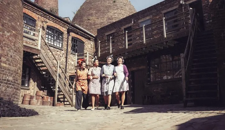 Four women in 1940s-style dresses walk through a brick courtyard with period industrial buildings and a kiln.