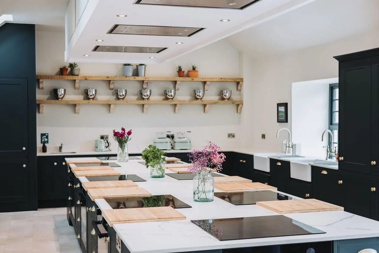 A modern cookery school kitchen with black cabinetry, white countertops, wooden shelving with plants, and multiple cooking stations along an island counter.