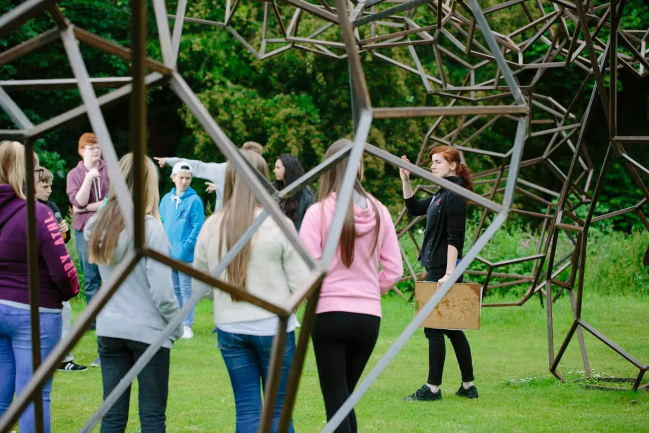 Group of visitors on a grassy field exploring a large sculptural art installation made of white metal branches.